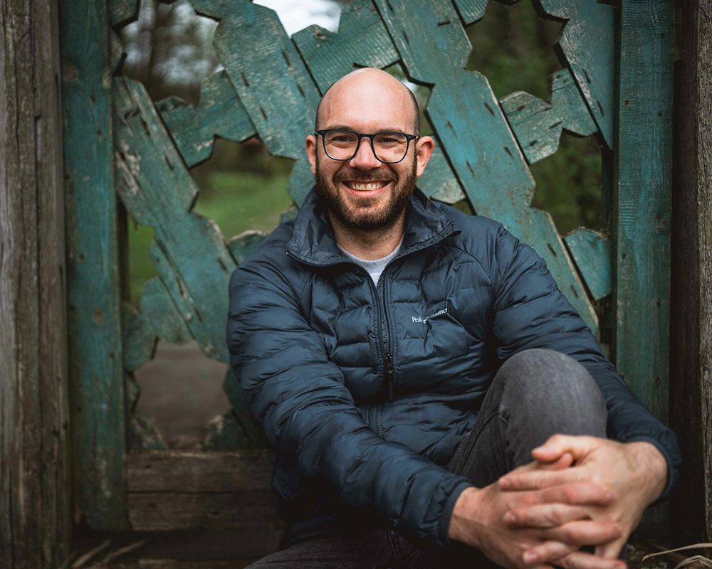 Ein Mann mit Glatze und Brille sitzt lächelnd im Freien. Er trägt eine dunkelblaue Pufferjacke und graue Hosen und sitzt vor einer verwitterten, dekorativen Holzkonstruktion mit einem blaugrünen geometrischen Muster. Der Hintergrund ist verschwommenes Grün und fängt den Charme ein, den jeder Hochzeitsfotograf Niederösterreichs bewundern würde.