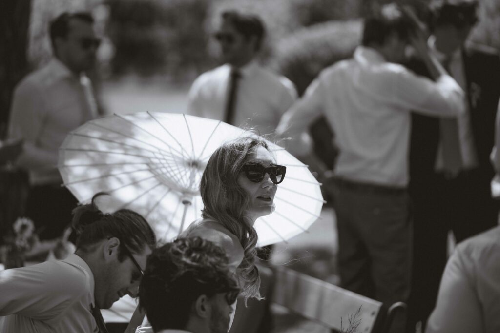 Ein Schwarzweißfoto zeigt eine Frau, die mit Sonnenbrille unter einem Sonnenschirm sitzt. Sie ist bei einer Veranstaltung im Freien von Menschen in formeller Kleidung umgeben. Einige stehen, andere sitzen auf Bänken unter Bäumen.