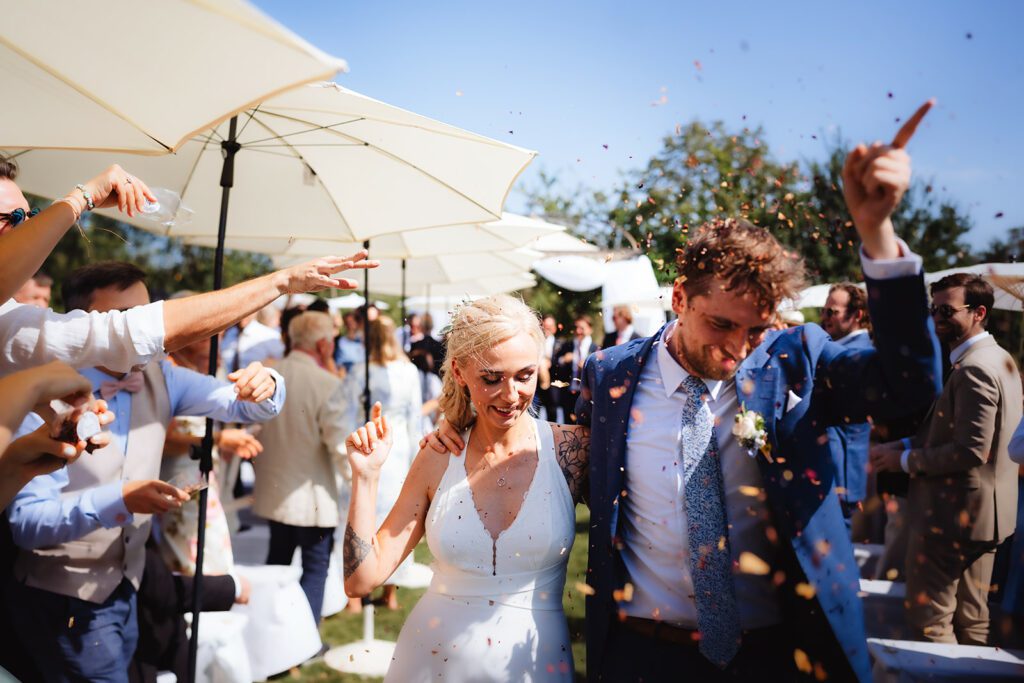 Ein fröhliches Paar im Hochzeitskleid geht Hand in Hand unter einem Konfettiregen hindurch. Sie sind an einem sonnigen Tag von feiernden Gästen unter weißen Sonnenschirmen umgeben und fangen einen festlichen Hochzeitsmoment im Freien ein.