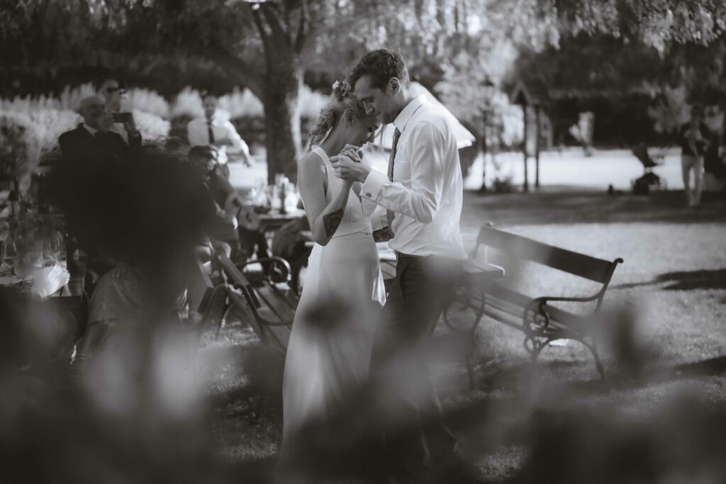 Ein Schwarzweißfoto eines Paares, das eng beieinander in einem Garten tanzt. Die Szene ist intim und ruhig, mit Bäumen im Hintergrund und einer Bank in der Nähe. In der Ferne sind ein paar Personen zu sehen, leicht verschwommen.