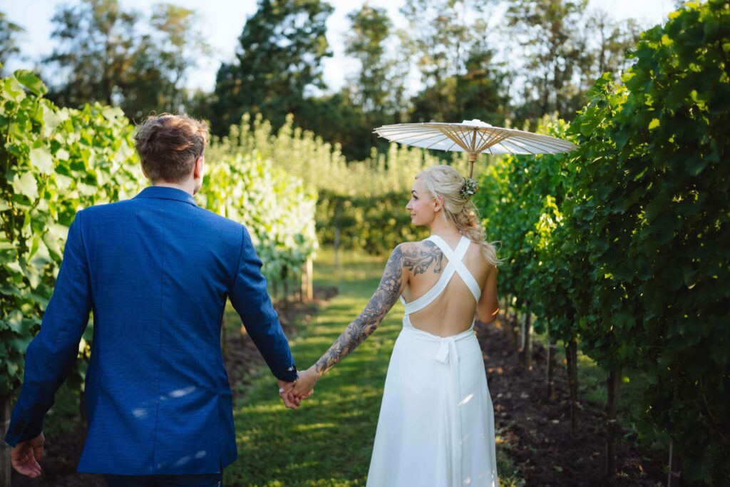 Ein Paar, wobei die Frau einen Sonnenschirm hält, geht Hand in Hand durch einen Weinberg. Der Mann trägt einen blauen Anzug und die Frau ein weißes Kleid mit sichtbaren Tattoos. Der Weinberg ist grün und üppig unter einem klaren Himmel.