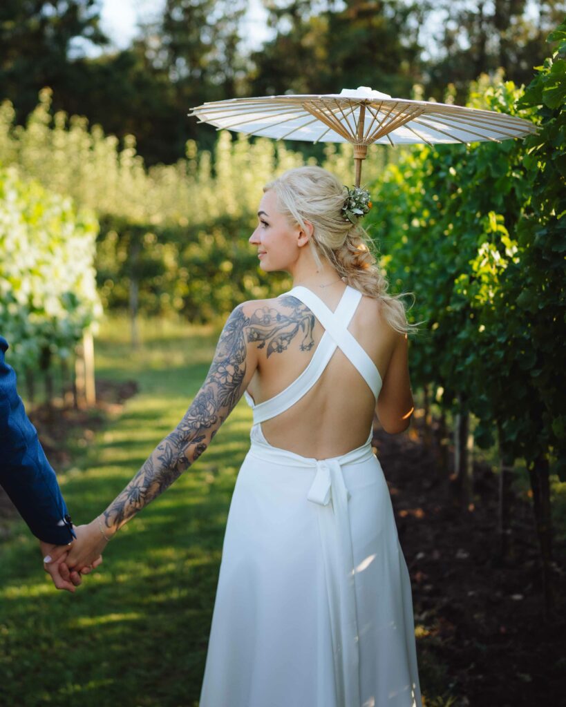 Im Herzen eines Weinbergs, aufgenommen von einem talentierten Hochzeitsfotografen aus Niederösterreich, steht eine Braut mit blondem Haar und tätowiertem Arm anmutig. Sie hält einen weißen Sonnenschirm in ihrem rückenfreien Kleid, Hand in Hand mit ihrem Partner, umgeben von leuchtend grünen Weinreben unter dem sonnigen Himmel.