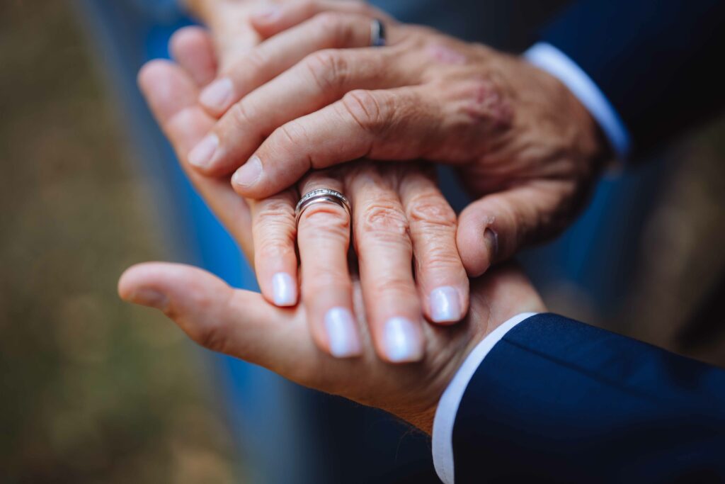 Nahaufnahme von zwei Personen, die sich zärtlich an den Händen halten. Die obere Person trägt einen silbernen Ring und hellen Nagellack, während die andere einen dunklen Anzug trägt. Der Hintergrund ist leicht verschwommen, was eine Szene im Freien vermuten lässt.