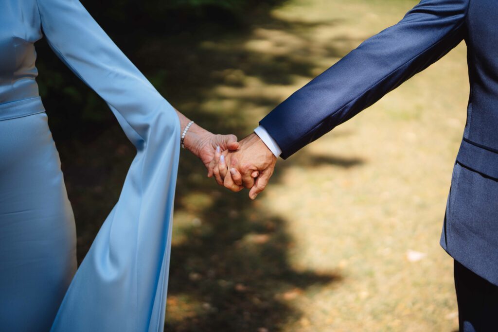 Ein älteres Paar steht Händchen haltend im Freien. Die Frau trägt ein langärmeliges, hellblaues Kleid und der Mann einen dunklen Anzug. Sonnenlicht erzeugt ein sanftes Spiel aus Licht und Schatten auf dem Boden.