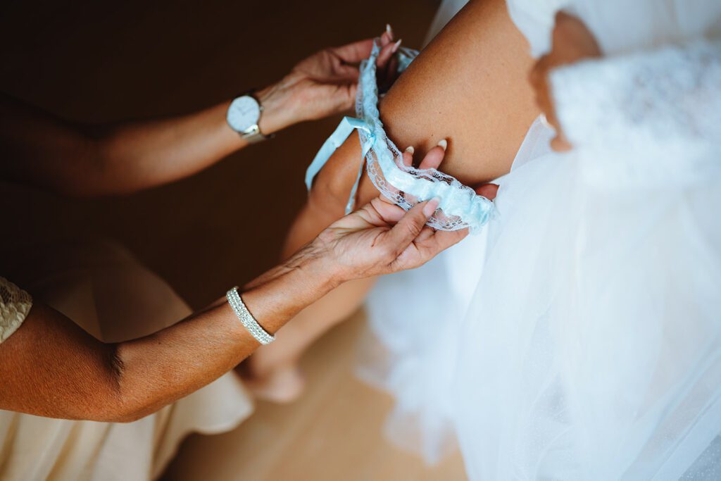 Eine Nahaufnahme von Sabrina, der Braut, in einem weißen Hochzeitskleid, während Michael ihr hilft, ein hellblaues Strumpfband um ihren Oberschenkel zu legen. Ihr Arm ruht sanft auf ihrem Bein und vermittelt ein Gefühl von Zärtlichkeit und Vorfreude.