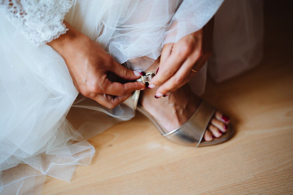 Sabrina, in einem weißen Kleid, schließt den Verschluss einer goldenen Sandale an ihrem Fuß. Der Holzboden ergänzt den durchsichtigen und zarten Stoff ihres Kleides, während ihre Nägel mit einer perfekten Schicht rosa Lack glänzen.