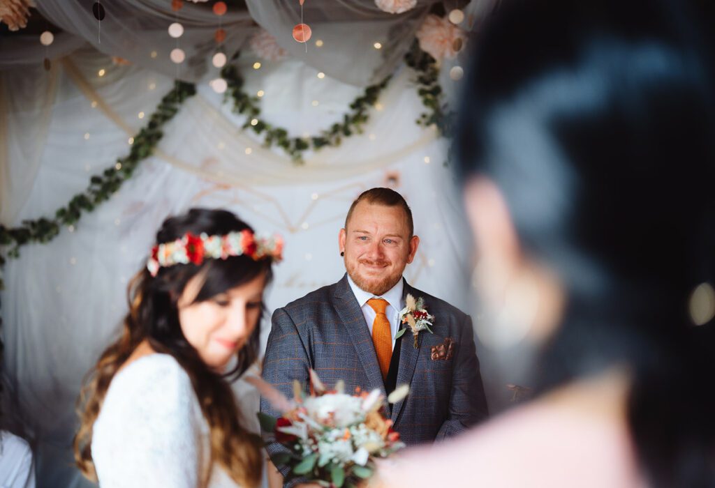 Sabrina und Michael stehen einander gegenüber. Er trägt einen Anzug und lächelt herzlich, während sie einen Blumenkranz trägt. Der Hintergrund ist mit drapiertem Stoff, Grünpflanzen und Lichtern geschmückt, die für eine festliche Note sorgen. Im Vordergrund ist teilweise eine weitere Person zu sehen, was den Charme der Szene noch verstärkt.