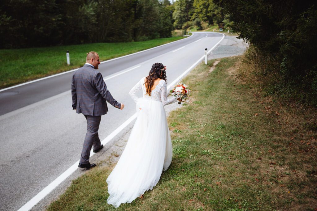 Eine Braut namens Sabrina in einem weißen Kleid mit Blumen in der Hand und ihr Bräutigam Michael in einem Anzug gehen eine kurvenreiche Straße entlang, die von Gras und Bäumen gesäumt ist. Die Szene hat eine friedliche, natürliche Kulisse mit viel Grün und einer kurvenreichen Straße, die sich vor ihnen erstreckt.