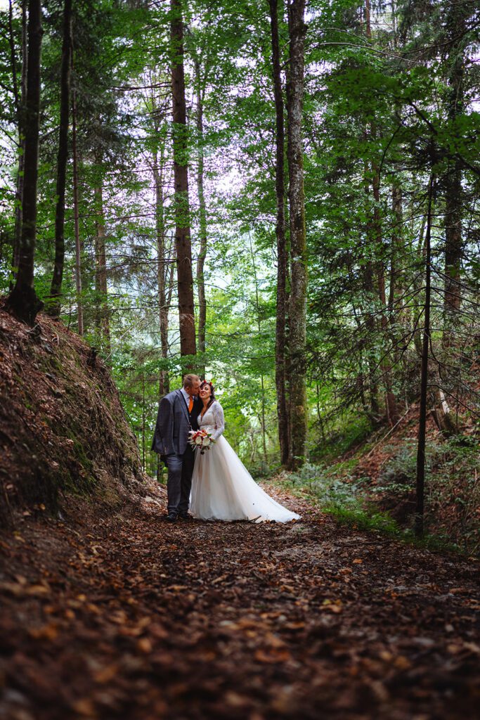 Sabrina und Michael stehen in ihrem Hochzeitskleid eng beieinander inmitten eines ruhigen Waldes. Die Braut, Sabrina, glänzt in einem weißen Kleid, während Michael in seinem Anzug elegant aussieht. Sie sind von hohen Bäumen und üppigem Grün umgeben, der Boden ist mit Blättern bedeckt.