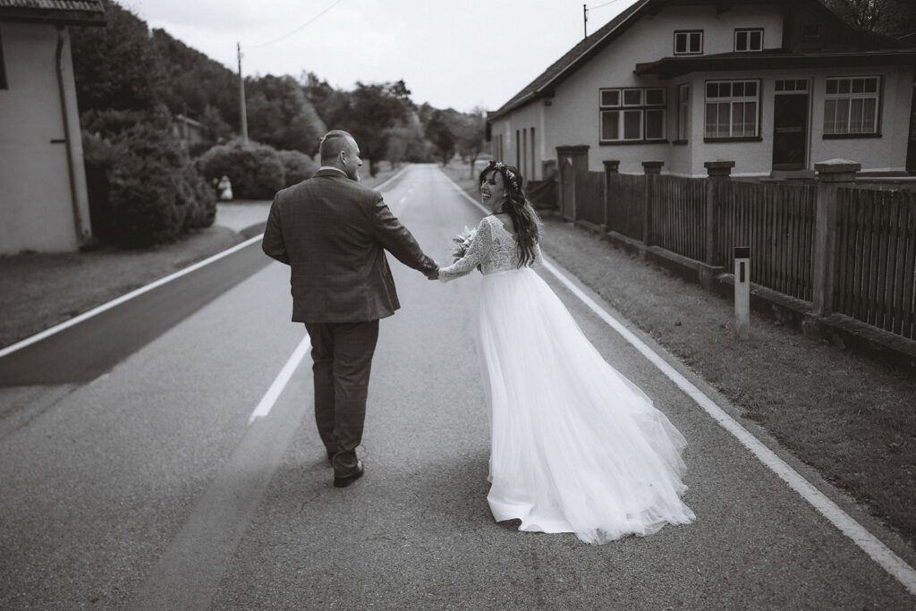 Braut und Bräutigam gehen Hand in Hand eine verlassene Straße entlang. Die Braut trägt ein langes weißes Kleid und der Bräutigam einen Anzug. Sie lächeln sich an, umgeben von einer friedlichen ländlichen Umgebung mit Häusern und viel Grün.