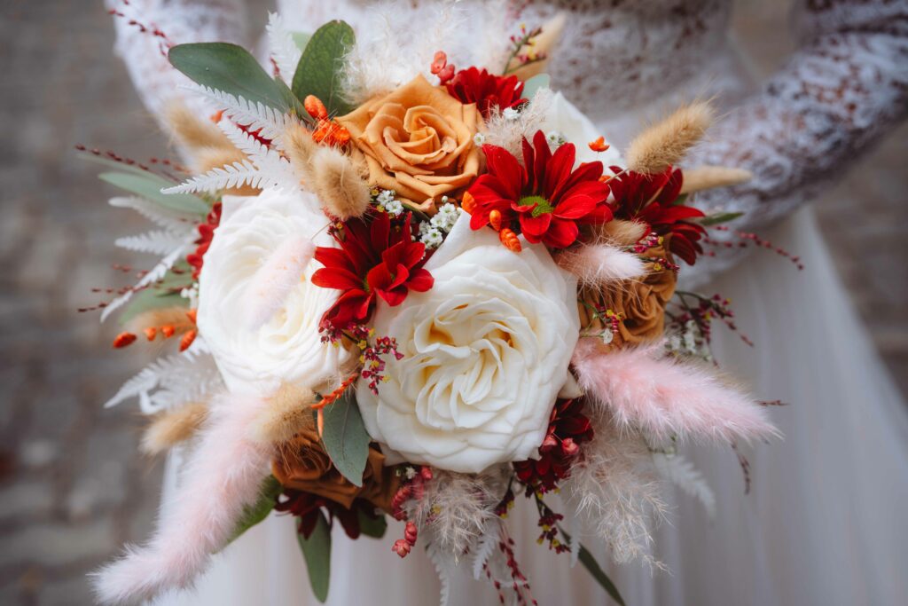 Eine Braut hält einen Strauß mit weißen und orangefarbenen Rosen, roten Gänseblümchen, grünen Blättern und zartrosa Pampasgras vor einem Kopfsteinpflasterhintergrund.