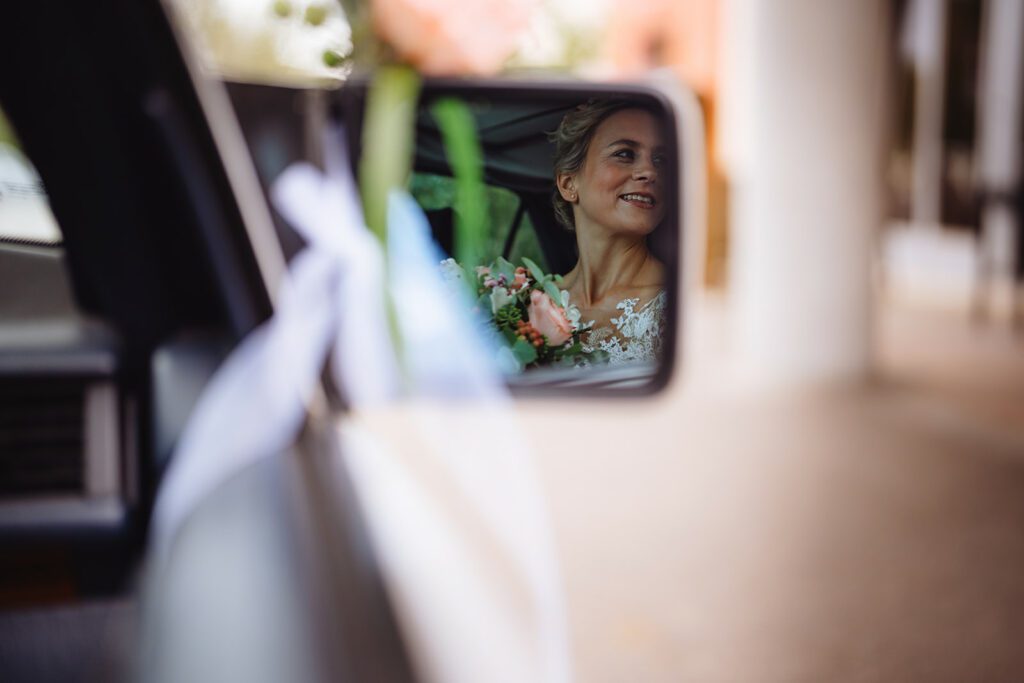 Im Seitenspiegel eines Autos sieht man eine lächelnde Frau im Brautkleid. Sie hält einen Blumenstrauß in der Hand und der Innenraum des Autos ist leicht zu erkennen. Der Hintergrund außerhalb des Autos ist unscharf.