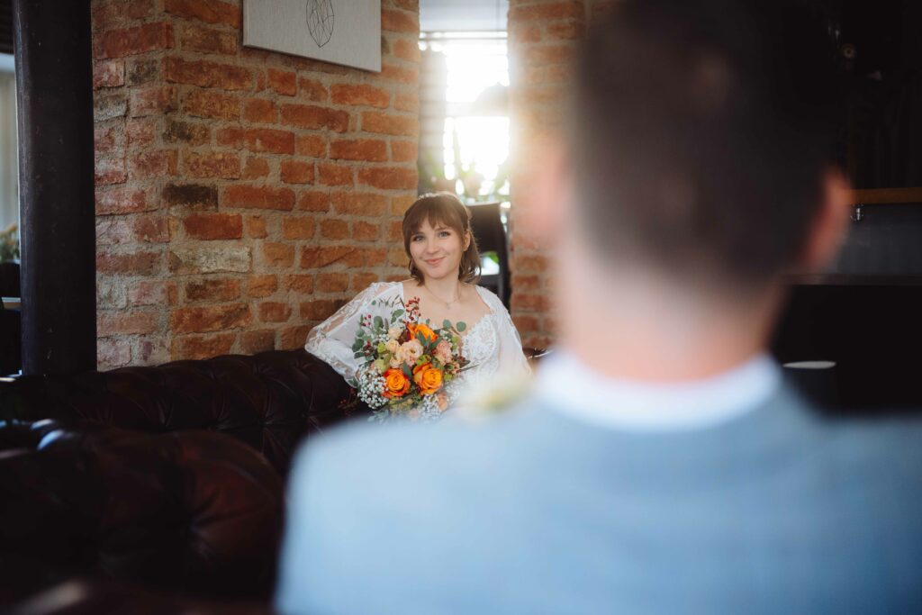 Eine Braut in einem weißen Kleid hält einen farbenfrohen Strauß orangefarbener und gelber Blumen in der Hand und sitzt auf einem Ledersofa in einem warm beleuchteten Raum mit Backsteinwänden. Im Vordergrund ist der Rücken einer Person im Anzug zu sehen.