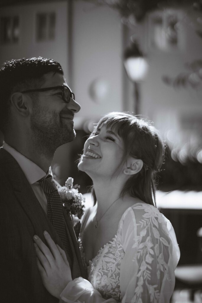 Ein Schwarzweißfoto, aufgenommen von einem Hochzeitsfotografen in Niederösterreich, zeigt ein lächelndes Paar an seinem Hochzeitstag. Die Braut im Spitzenkleid blickt freudig auf den Bräutigam mit Brille und Anzug. Sie stehen im Freien, mit sanft verschwommenen Gebäuden, die eine zauberhafte Kulisse bilden.