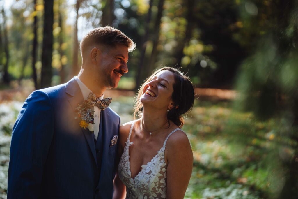 Ein Paar steht in einem sonnenbeschienenen Wald und lacht und lächelt einander an. Die Frau trägt ein weißes Spitzenkleid und der Mann einen blauen Anzug mit einer geblümten Fliege. Sonnenlicht fällt durch die Bäume und schafft eine warme, leuchtende Atmosphäre.