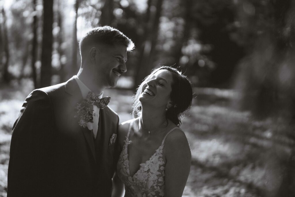 Ein Schwarzweißfoto eines lächelnden Paares in Hochzeitskleidung, das in einem sonnenbeschienenen Waldstück steht. Die Braut trägt ein Spitzenkleid und der Bräutigam einen Anzug mit Fliege. Beide genießen einen freudigen Moment zusammen.