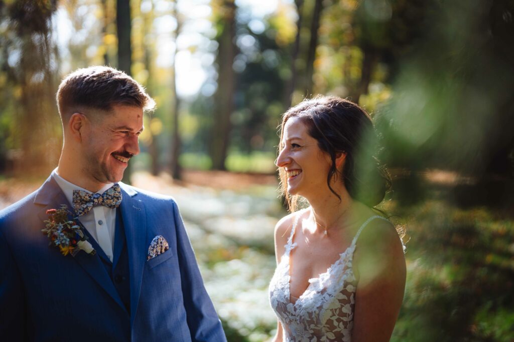 Braut und Bräutigam lächeln sich draußen in einem Wald an, während das Sonnenlicht durch die Bäume fällt. Der Bräutigam trägt einen blauen Anzug mit einer Blumenboutonniere, während die Braut ein Spitzenhochzeitskleid trägt.