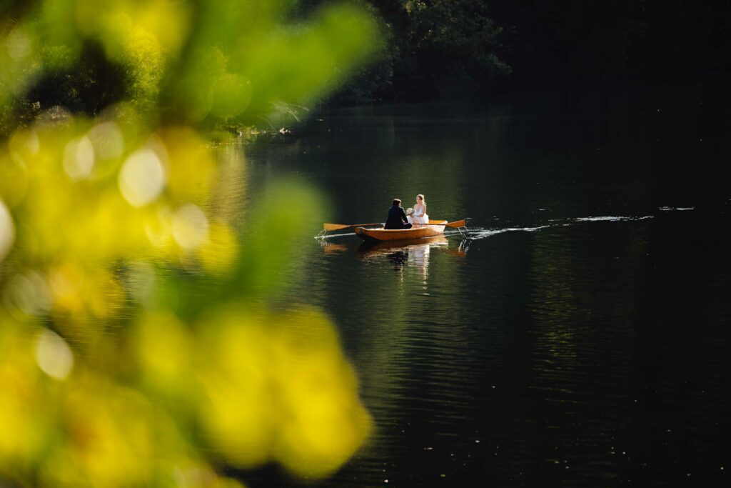 Ein Paar rudert in einem Holzboot auf einem ruhigen See, der von üppigem Grün umgeben ist. Sonnenlicht fällt durch die Blätter und schafft eine heitere und ruhige Atmosphäre.