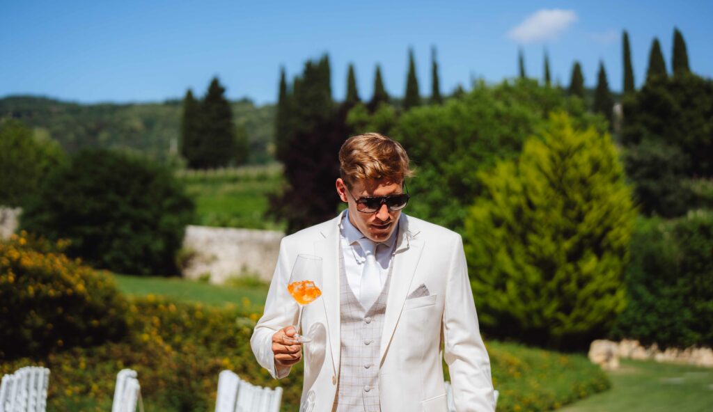 Ein Mann in weißem Anzug und Sonnenbrille hält ein Getränk mit einer Orangenscheibe in der Hand. Er steht im Freien, im Hintergrund sieht man üppiges Grün und Zypressen.