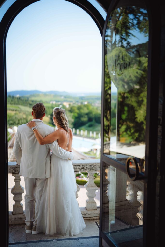 Ein Paar im Hochzeitskleid steht umarmt auf einem Balkon und blickt auf eine malerische Landschaft. Der Blick umfasst Grünflächen und einen fernen Horizont, eingerahmt von einem offenen Torbogen.