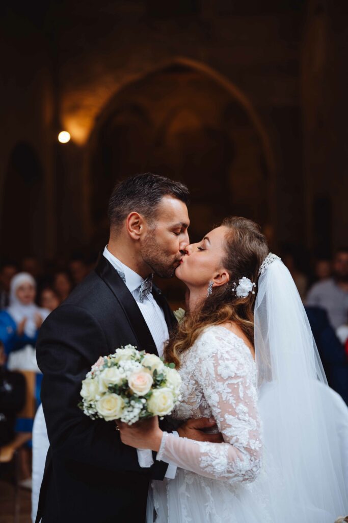 Braut und Bräutigam küssen sich in einem schwach beleuchteten Raum. Die Braut trägt ein weißes Spitzenhochzeitskleid mit Schleier und hält einen Strauß weißer und rötlicher Rosen in der Hand. Der Bräutigam trägt einen schwarzen Smoking. Die Gäste sind im Hintergrund verschwommen.