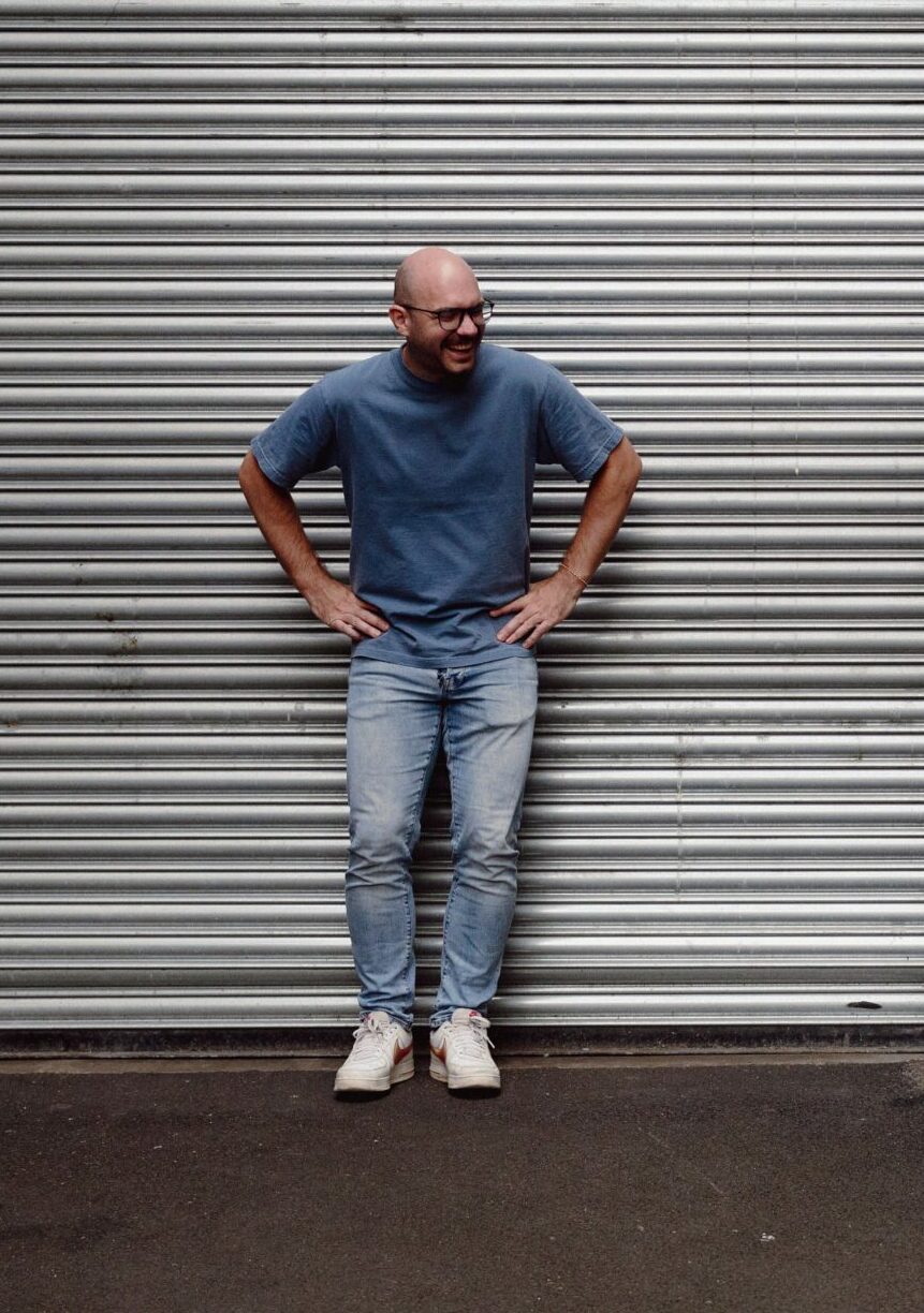 Ein kahlköpfiger Mann in blauem Hemd und Jeans steht mit den Händen in den Hüften vor einem Metallrollladen, während eine Frau mit langen Haaren in rosa Top und Jeans etwa ein paar Meter entfernt steht, lächelt und ein Telefon in der Hand hält. Beide tragen weiße Turnschuhe.