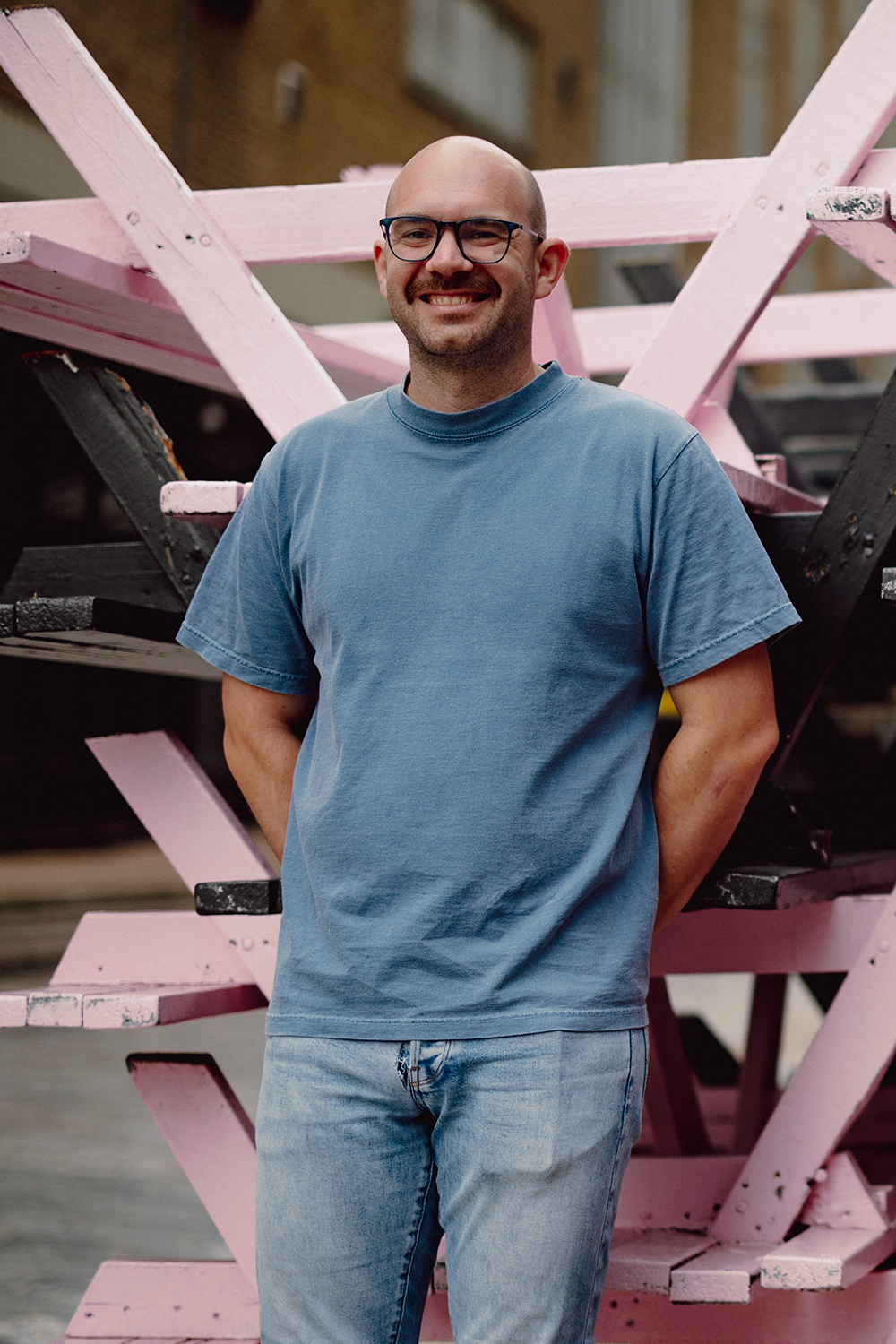 Eine Person mit Brille, Schnurrbart und Bart steht lächelnd vor einer abstrakten Struktur aus ineinandergreifenden rosa und schwarzen Holzbalken. Sie trägt ein blaues T-Shirt und hellblaue Jeans. Der Hintergrund ist eine städtische Außenkulisse.