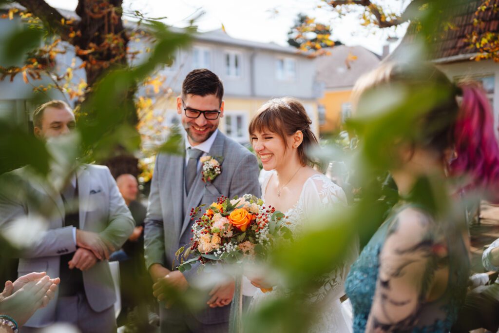 Braut und Bräutigam stehen zusammen und lächeln freudig. Die Braut hält einen bunten Blumenstrauß und sie sind von ein paar Menschen in einer malerischen Umgebung im Freien mit elegantem Grün und Sonnenlicht umgeben. Dieser bezaubernde Moment könnte leicht von einem talentierten Hochzeitsfotografen aus Österreich festgehalten werden.