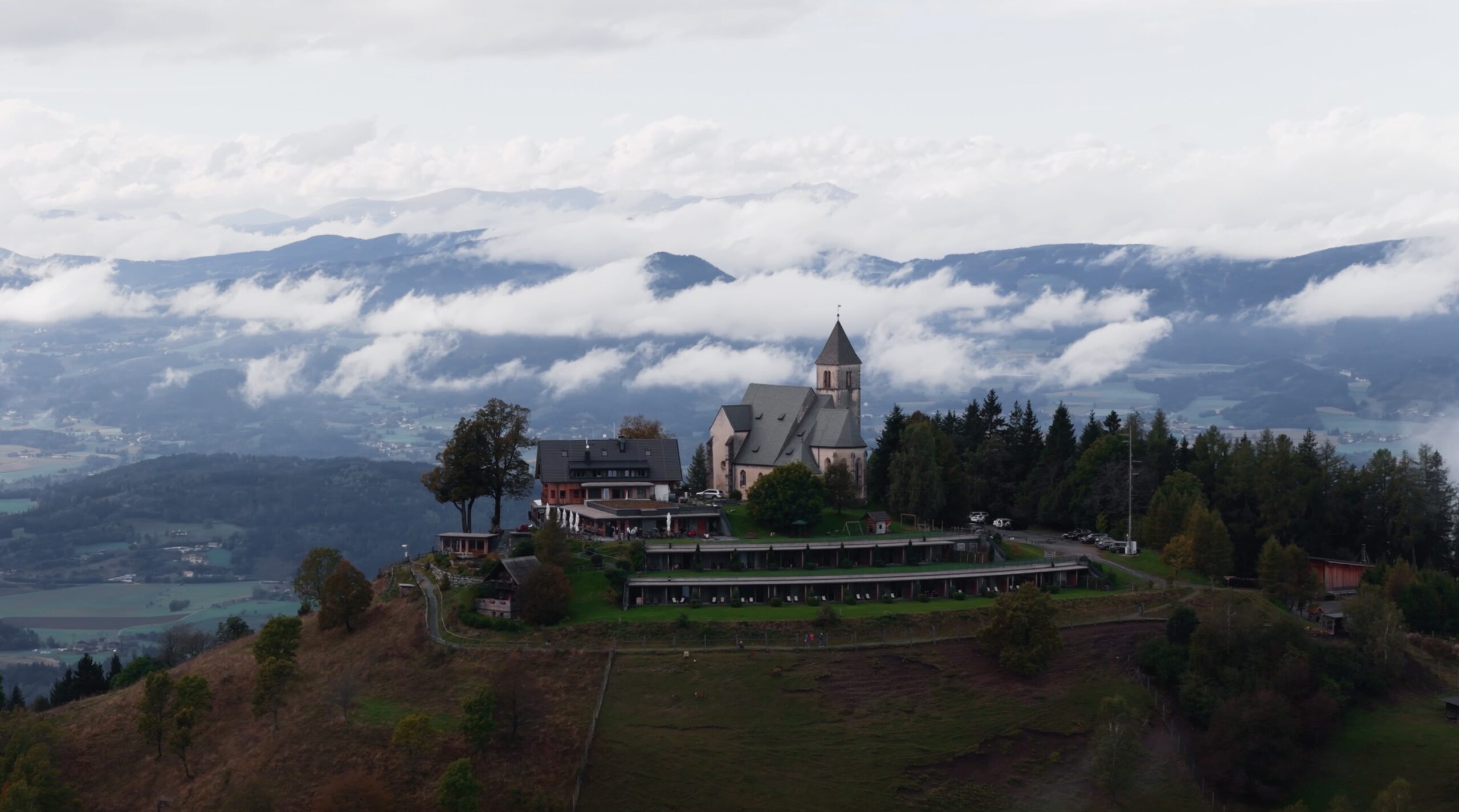 Eine bezaubernde Kirche steht auf einem Hügel in Niederösterreich, umgeben von Bäumen, perfekt für eine Hochzeit. In der Nähe ergänzt ein langes Gebäude die ruhige Landschaft. Unter ihnen sind Täler in tief hängende Wolken gehüllt und Berggipfel tauchen unter einem wolkigen Himmel durch den Nebel auf.