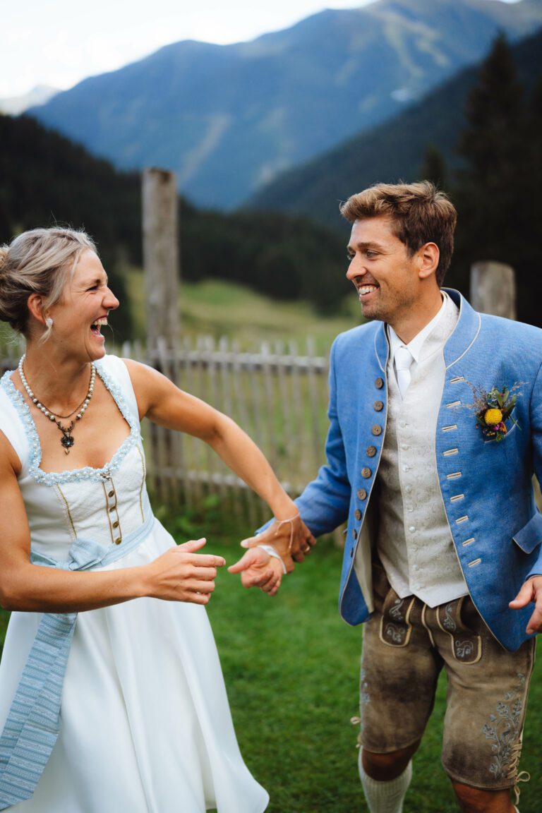 Ein fröhliches Paar in traditioneller bayerischer Kleidung steht draußen für seine Hochzeitsfotos in Salzburg. Die Frau trägt ein weißes Kleid und der Mann eine blaue Jacke und Lederhosen. Sie halten Händchen, lächeln inmitten der Berge und eines Holzzauns und fangen die Essenz einer emotionalen Hochzeit ein.