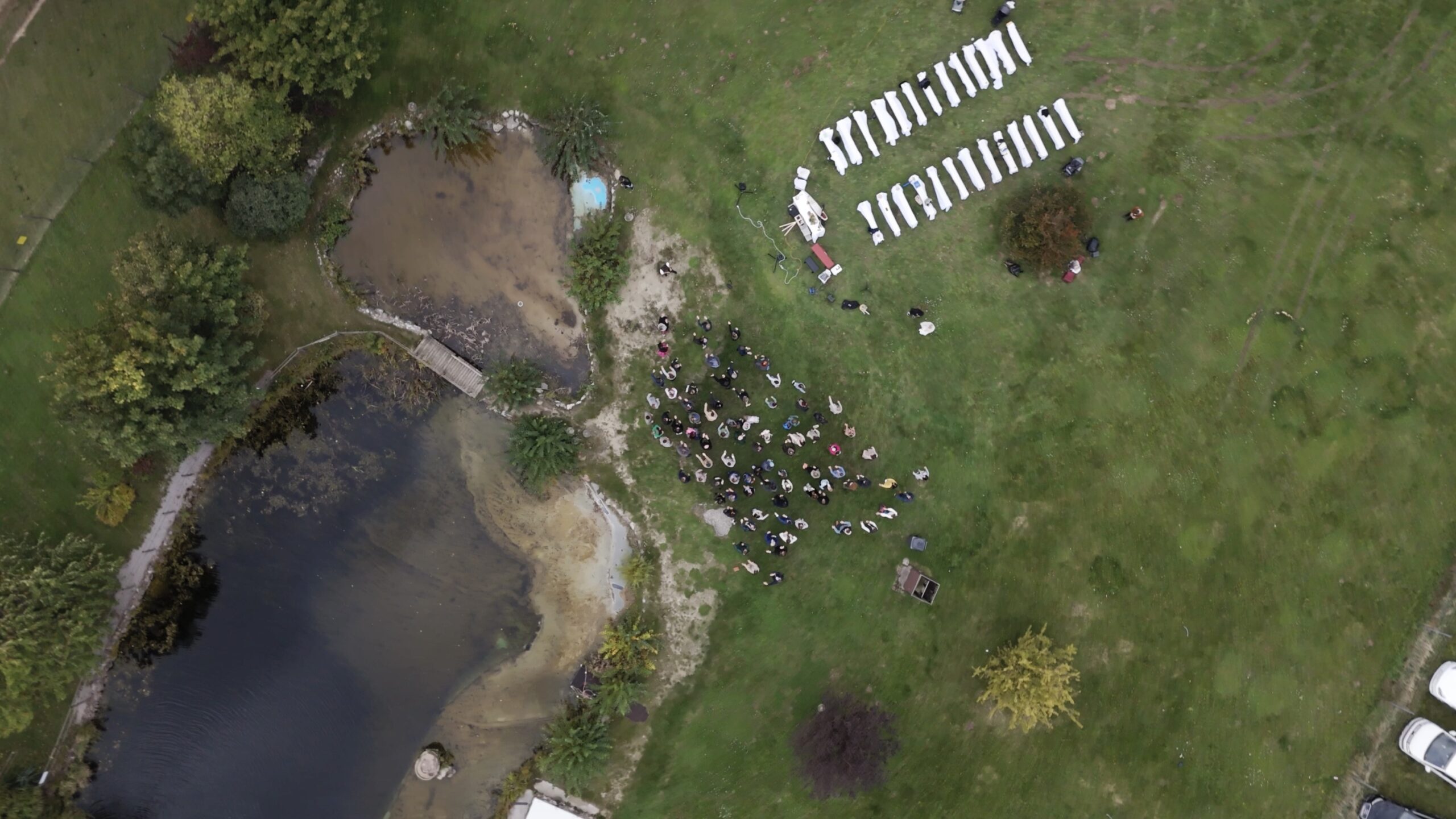 Luftaufnahme einer Hochzeitszeremonie. Reihen weißer Stühle zieren den grünen Rasen neben einem kleinen Teich und fangen die perfekte Atmosphäre für einen Hochzeitsfilm Kärnten ein. Die Gäste versammeln sich zwischen den Stühlen und dem Teich, was auf eine Zeremonie im Freien hindeutet, die von den umliegenden Bäumen eingerahmt wird.